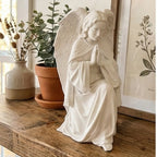 White angel statue on a wooden surface with plants and a framed picture in the background