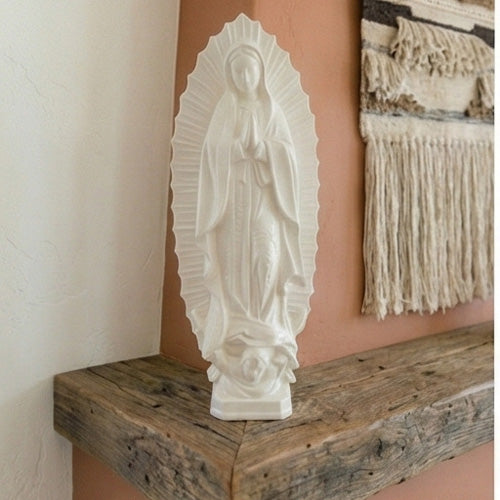 White Our Lady of Guadalupe Sculpture on a wooden shelf against a wall with a textured painting.